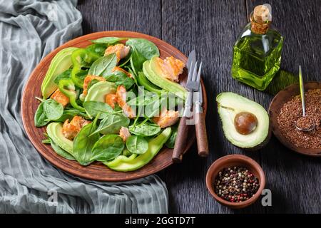 Lachs Spinat Avocado und grüner Pfeffer Salat auf einem braunen rustikalen Teller mit Gabel und Messer auf einem Mooreichentisch, mediterrane gesunde Ernährung Stockfoto