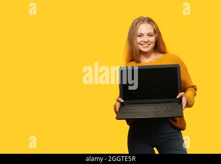 Blonde Frau hält Laptop mit leerem Bildschirm auf gelbem Hintergrund. Speicherplatz kopieren. Frau hält den Laptop-Bildschirm mit einem Modell nach vorne. Stockfoto