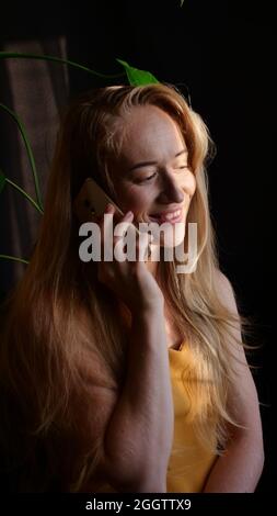 Eine junge Frau, ein blondes kaukasisches Mädchen, spricht an einem sonnigen Tag am Telefon und lächelt, während sie sich zu Hause isoliert. Rufen Sie den Service an der Fernarbeit an. Stockfoto