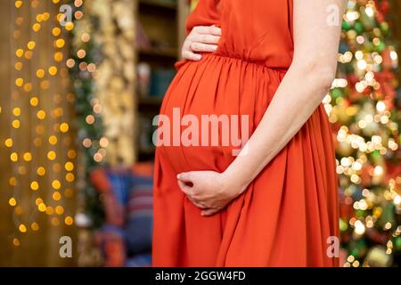 Bestes weihnachtsgeschenk. Gesichtslose Schwangere in einem roten Kleid umarmt ihren Bauch vor dem Hintergrund des Weihnachtswohnzimmers in Unschärfe. Nahaufnahme kein fa Stockfoto