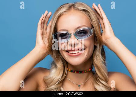 Fröhliche junge Frau in trendigen Sonnenbrillen lächelt isoliert auf blau Stockfoto