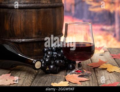 Glas Rotwein mit Trauben, Weinflasche und Fass auf Holztisch im Freien mit Herbstblättern und Hintergrund Stockfoto