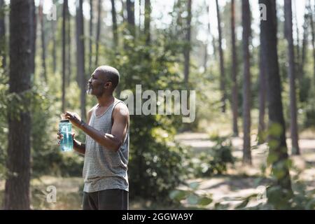 Seitenansicht eines älteren afroamerikanischen Sportlers, der im Wald eine Sportflasche hält Stockfoto