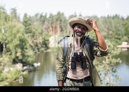 Positiver afroamerikanischer Wanderer mit Hut und Fernglas, der im Freien auf die Kamera schaut Stockfoto