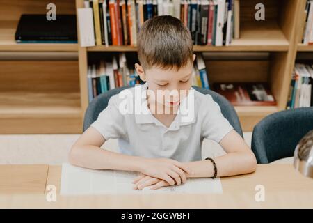 Schuljunge sitzt auf dem Tisch und liest Buch in der Bibliothek in der Schule. Vorbereitung auf Hausaufgaben. Ein guter Student. Der Junge liebt es zu lesen. Freier Platz am sch Stockfoto