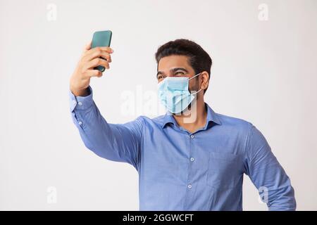 Porträt eines Mannes, der auf Selfie im Handy klickt, mit Maske im Gesicht vor einfachem Hintergrund. Stockfoto