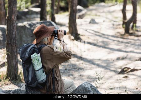 Ältere Reisende mit Rucksack und Karte, die durch ein Fernglas im Wald schaut Stockfoto