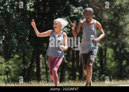 Glückliches multiethnisches Paar mit Sportflasche, die mit den Fingern zeigt, während sie im Park läuft Stockfoto