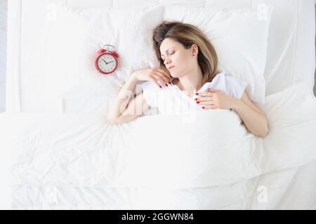 Junge Frau, die im Bett in der Nähe des roten Weckers liegt, Draufsicht Stockfoto