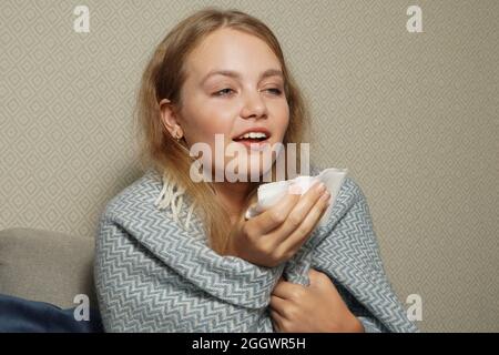 Frau niest in weißem Gewebe. Junge blonde Frau, in warme Decke gehüllt, niest, hat Grippe. Stockfoto