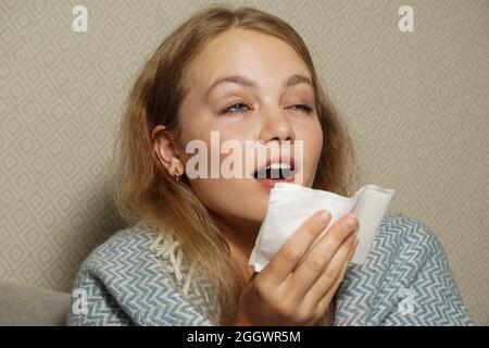 Frau niest in weißem Gewebe. Junge blonde Frau, in warme Decke gehüllt, niest, hat Grippe. Stockfoto