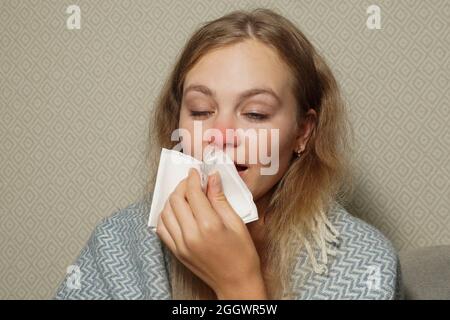 Frau niest in weißem Gewebe. Junge blonde Frau, in warme Decke gehüllt, niest, hat Grippe. Stockfoto