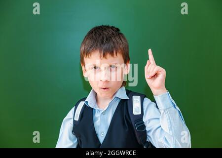 Lustige Schuljunge Grimassen in der Nähe der grünen Schultafel im Klassenzimmer. Grundschulkind mit Tasche. Zurück zur Schule. Stockfoto