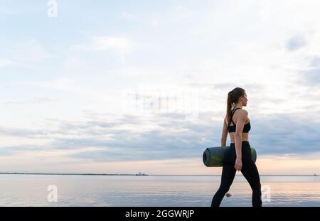 Das junge Fitnessmodell mit Yogamatte lässt bei Sonnenuntergang gegen den Himmel Stockfoto