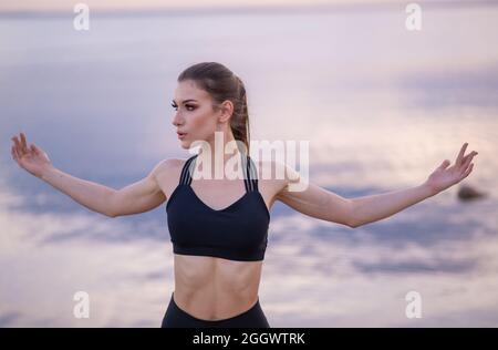 Junges schönes Mädchen, das sich aufwärmt, bevor es Yoga am Meer bei Sonnenuntergang macht Stockfoto