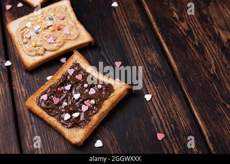 Toast mit Schokoladenpaste und Erdnussbutter, bestreut mit bunten Herzen auf dunklem Holzhintergrund, süßes Dessert. Stockfoto