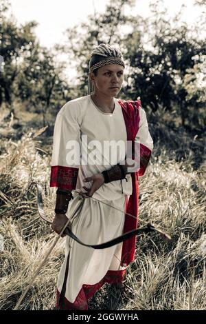 Die antike griechische Bogenschützin in weißem Kleid steht zwischen der Wiese mit Pfeil und Bogen in den Händen. Stockfoto