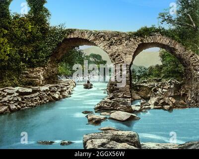 Ein Blick aus dem frühen 20. Jahrhundert auf einen Bootsführer, der die Old Weir Bridge im Killarney National Park in der Grafschaft Kerry, Irland, passieren wird. Das genaue Alter der Alten Weirbrücke ist unklar, aber es wird angenommen, dass sie bis zum 16. Jahrhundert über die schnell fließenden Gewässer und Strömungen an dieser Stelle überquert wurde. Seine einzigartige Lage beim Meeting of the Waters der drei Seen von Killarney sorgt dafür, dass es seit Hunderten von Jahren ein beliebtes Reiseziel ist. Stockfoto