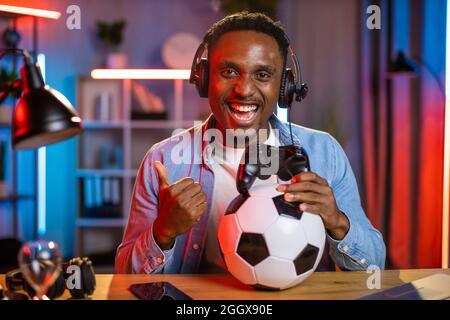 Fröhlicher junger Mann mit Kopfhörern, der den Daumen auf die Kamera zeigt, während er mit Joystick und Fußball am Tisch sitzt. Glücklich schwarzen Kerl verbringen Freizeit zu Hause für das Spielen von Videospielen. Stockfoto
