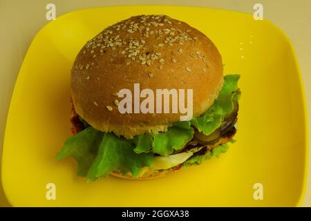 Auf einer gelben quadratischen Keramikplatte liegt ein großes Sandwich. Der Teller wurde aus nächster Nähe auf einem beigefarbenen Holztisch geschossen Stockfoto
