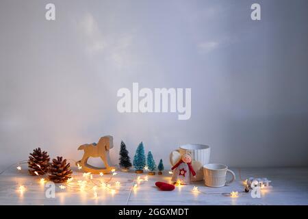 Weihnachtskomposition auf dem Tisch. Graues Schaukelpferd aus Holz, vier kleine künstliche schneebedeckte Bäume, Engel, Girlande und Becher Stockfoto