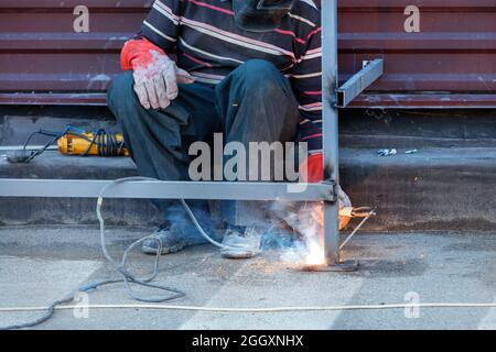 Ein heller Lichtbogen, während ein sitzender Schweißer einen Metallzaun um das Haus herum installiert und verstärkt. Speicherplatz kopieren. Stockfoto