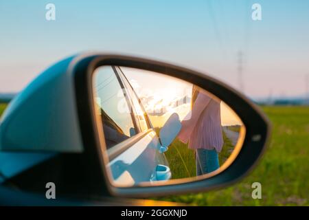 Rückspiegel mit einer Spiegelung der weiblichen Silhouette lädt ein Elektroauto, im Hintergrund sichtbaren Himmel bei Sonnenuntergang Stockfoto