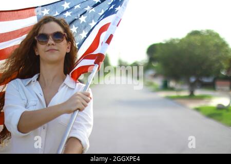 Junge Rotschopf hispanische und kaukasische Frau trägt Sonnenbrille winkt die amerikanische Flagge draußen. Stockfoto