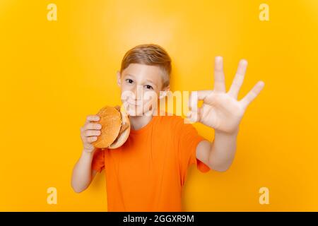 Ein Junge isst einen Hamburger und zeigt auf gelbem Hintergrund OK Stockfoto