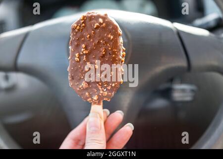 Hand hält eine Eiscreme mit Schokoladenmandelnüssen auf dem Stock mit Hintergrund des Lenkrads auf der Fahrt Stockfoto