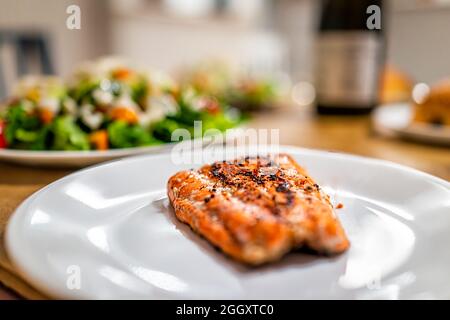 Gegrillter Sockeye-Lachs, Meeresfrüchte, Fisch aus der Nähe auf weißem Teller mit schwarzer Pfefferwürze und verschwommenem Hintergrund mit Salat und Wein zum Abendessen Stockfoto