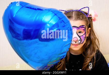 Kind mit Gesichtsbemalung von Kitty hält Folienballon, nettes kleines Mädchen mit bemalten bunten Maske der Katze auf ihrem Gesicht. Porträt eines schweren Kindes mit Ba Stockfoto