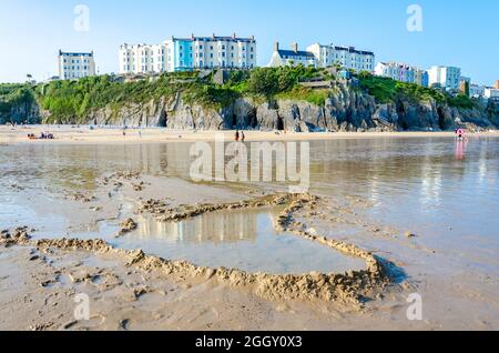 Ein Staudamm, der bei Ebbe am Tenby South Beach errichtet wurde, hat einen Wasserpool geschaffen. Dahinter befinden sich farbenfrohe Gebäude auf der Esplanade auf der Klippe darüber. Stockfoto