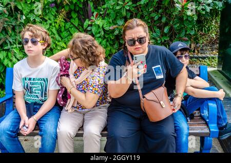 Eine Familie sitzt zusammen auf einer Parkbank, während Mama einen Videoanruf auf ihrem Mobiltelefon entgegennimmt. Stockfoto