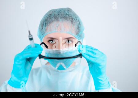 Eine Krankenschwester in einem medizinischen Kleid, einer Maske und Schutzhandschuhen trägt eine transparente Brille, die eine Spritze hält Stockfoto