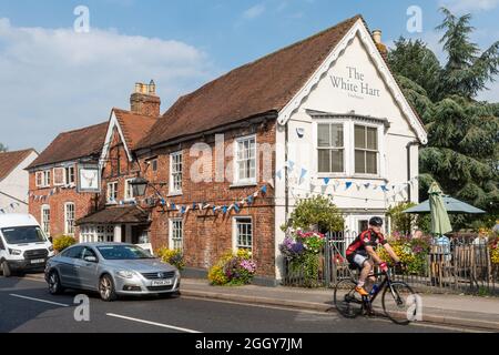 The White Hart Pub an der High Street in Chobham Village, Surrey, England, UK, im Sommer mit einem Radfahrer und Verkehr Stockfoto