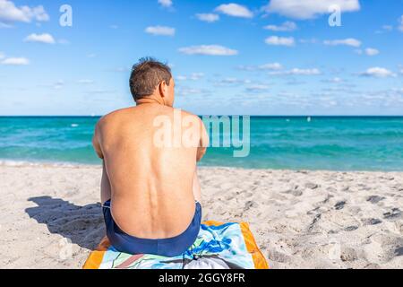 Türkisfarbenes, farbenprächtiges Wasser und ein brauner Mann, der auf einem Handtuch sitzt und während der sonnigen Sonne auf die Sandküste im Sunny Isles Beach, North Miami, Florida blickt Stockfoto