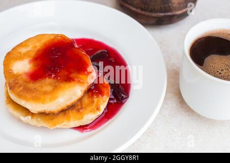 Frische Käsekuchen mit Pflaumenmarmelade und eine Tasse mit frisch gebrühtem Kaffee auf einer leichten Tischplatte. Wachen Sie morgens auf, beginnen Sie einen neuen Tag. Herzhaftes Frühstück. Stockfoto