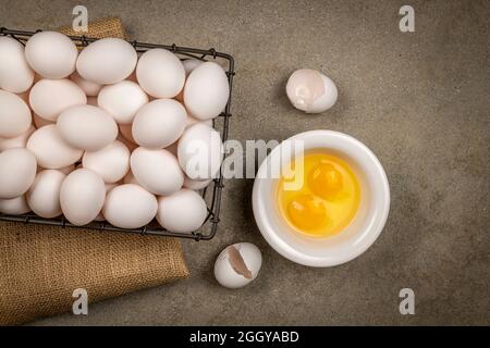 Ein kleiner Drahtkorb mit frischen Eiern mit zwei zerrissenen Eiern und Eigelb ruht auf einer Zementplatte mit Heu, während das Frühstück vorbereitet wird. Stockfoto