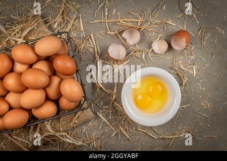 Ein kleiner Drahtkorb aus frischen braunen Eiern mit zwei zerrissenen Eiern und Eigelb ruht auf einer Zementplatte mit Heu, während das Frühstück vorbereitet wird. Stockfoto