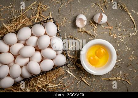 Ein kleiner Drahtkorb mit frischen Eiern mit zwei zerrissenen Eiern und Eigelb ruht auf einer Zementplatte mit Heu, während das Frühstück vorbereitet wird. Stockfoto