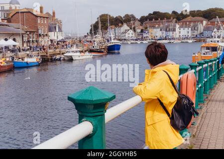 Seitenansicht Frau im gelben Regenmantel, die den Blick auf die Bucht mit Schiffen und Booten der Küstenstadt genießt. Reisen vor Ort. Weymouth, England Stockfoto