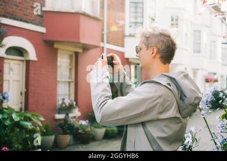 Junger männlicher Tourist, der während eines Spaziergandes in der Wohnsiedlung Cottagecore Video dreht oder Fotos auf dem Mobiltelefon fotografiert. Wunderschöner Blick auf die Straße Stockfoto