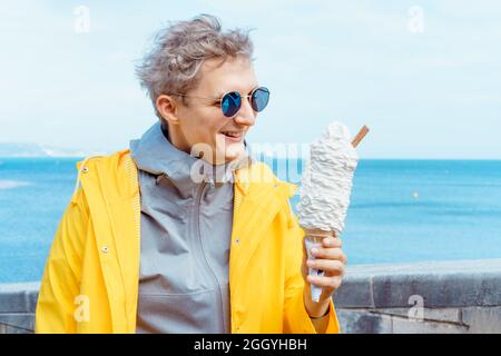Aufgeregt kaukasischen jungen männlichen Touristen in dunklen Sonnenbrillen und gelben Regenmantel essen riesige Eis in einem Waffelkegel während eines Spaziergangs in der Nähe des Meeres Stockfoto