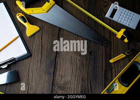 Thema des Auftragnehmers. Werkzeugsatz des Auftragnehmers: Gelber Harthut, libella, Handsäge. Pläne und Notizbuch auf dem rustikalen braunen Plankenhintergrund. Stockfoto