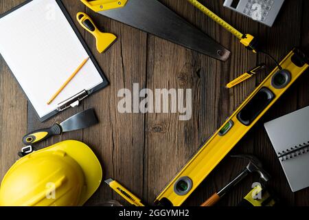 Thema des Auftragnehmers. Werkzeugsatz des Auftragnehmers: Gelber Harthut, libella, Handsäge. Pläne und Notizbuch auf dem rustikalen braunen Plankenhintergrund. Stockfoto