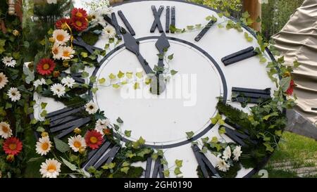 Große Wanduhr auf dem Feld in blühenden Blumen, zeigt die Zeit 11:55, 12:00. Zeitmanagement-Konzept, Sommerzeit. Frühling oder Hochzeit Stockfoto