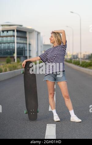 Erwachsene Frauen laufen gerne auf Longboard auf einer leeren Stadtstraße. Glückliche fröhliche Frau hält Skateboard Stockfoto