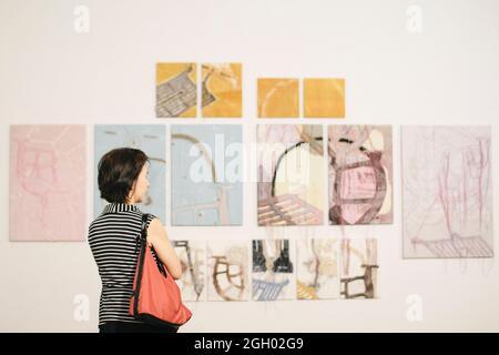 Asiatische Frau, die in einer Kunstgalerie vor farbenfroh gerahmten Gemälden auf einer weißen Wand steht Stockfoto