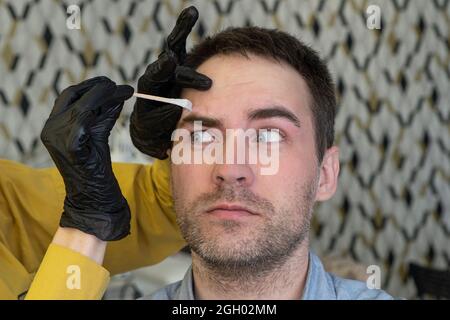 Kosmetikerin, die im Schönheitssalon wunderschöne bärtige Männerbrauen mit einer Pinzette zupft. Weibliche Hände pflücken Augenbrauen Pinzette zu entspannten bärtigen brutalen Mann Stockfoto